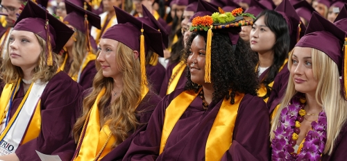 Cronkite School graduates sit in a line wearing their graduation gowns, hats and stoles.