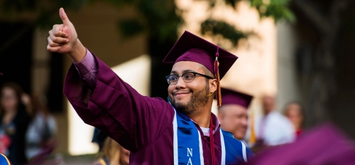 A man in a graduation gown with a Navy military stole smiles and gives a thumbs up to the crowd