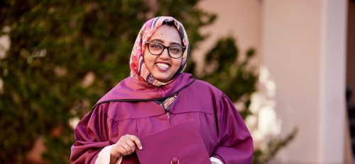 Woman in a graduation cap and gown, head scarf and glasses, smiling widely.