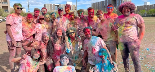 A group of people covered in colorful splashes of powder pose outside.