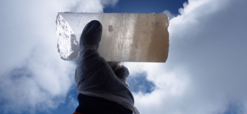 Gloved hand holding an ice core from Antarctica up to a cloud-filled blue sky.