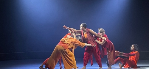 Dancers in bright yellow and red clothing perform on a backlit blue and black stage. 