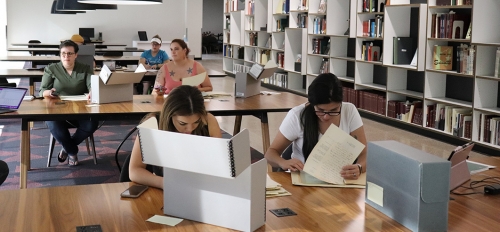 Students seated in a library at tables on which large containers are sprawled. Next to them are shelves of neatly stacked archival materials. The students are sorting through papers from the containers.
