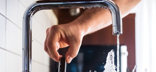 Hand turning on a faucet, out of which water pours into a glass.