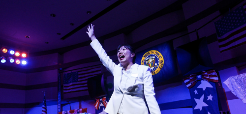 Performance artist, comedian and writer Kristina Wong stanidng on a stage with American flags, wearing a white suit, raising her arm and smiling.
