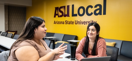 Two students seated in a classroom talking to each other. In the background, a sign on a wall says "ASU Local."