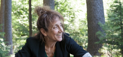 Liz Lerman sitting and looking to the side in a forest-like setting.