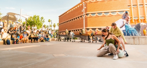 A crowd gathered outside of a circular building watches people perform a dance.