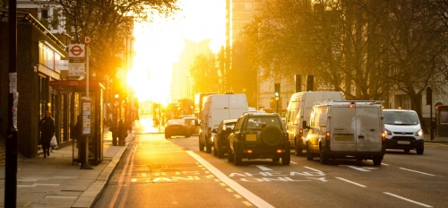 City street full of cars, surrounded by tall buildings and lit by blazing sunlight.
