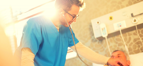 In this stock image a nurse is using his stethoscope to check the lung function of a patient in a hospital bed.