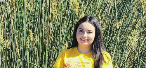 Odalis Amaya Amaya wears blue jeans and a yellow ASU T-shirt. Her arms are crossed in front of her. She is smiling and has long dark wavy hair. She is standing in front of a gray fence in front of tall grass.