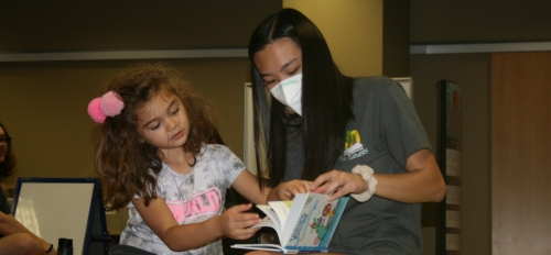 A college student wearing a mask read a book to a 7-year-old girl who is turning the pages