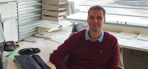 Petr Šulc smiling at the camera and sitting at an office desk with a computer and papers surrounding him.