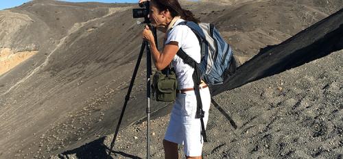 A dark-haired woman in white shorts and a white t shirt stands at a tripod on a gravel hill, taking a photograph