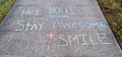 Sidewalk with chalk lettering that reads "Hey you! Stay awesome + smile."