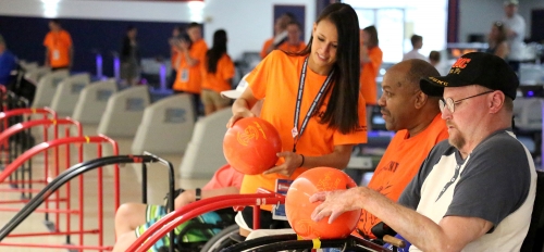 A woman holding a bowling ball speaks to two men in wheelchairs holding bowling balls.