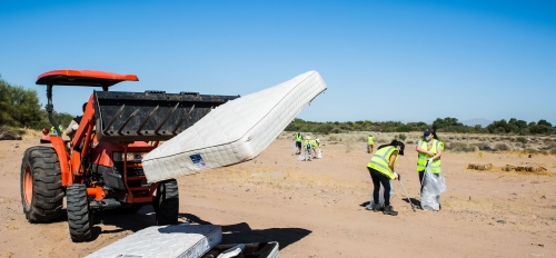 A forklift carries mattresses as volunteers in the background pick up trash on a dry riverbed