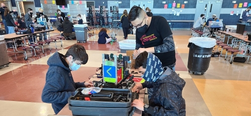 Three masked high school seniors work on a robot with four wheels at a table with tools strewn about