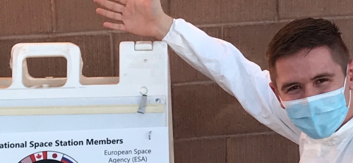 Eli Fox squats in front of a brick wall. He is wearing an all-white jumpsuit with a blue NASA logo over the breast. He has short brown hair and is wearing a blue surgical mask. His arm is extended to point at a sandwich board displaying scientific facts.
