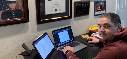 ASU student Ryan Johnston seated at a desk with a laptop. The wall behind the desk features photos and memorabilia from his time in the military.