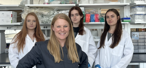 Candace Lewis and her lab assistants, standing behind her, posing for a photo in lab gear and in a lab setting.