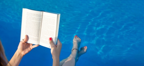 Hands holding a book above a pool in which a person's legs are dipped.
