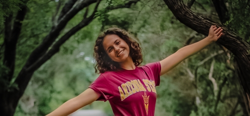 Sophia Godinez pictured smiling with arms and one leg raised in front of a dirt path surrounded by trees.