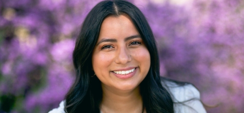 Portrait of Yazmin Reyes, a smiling young woman with dark hair and purple flowers in the background.