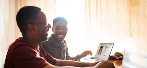 Two Black men smile as they work at laptops.