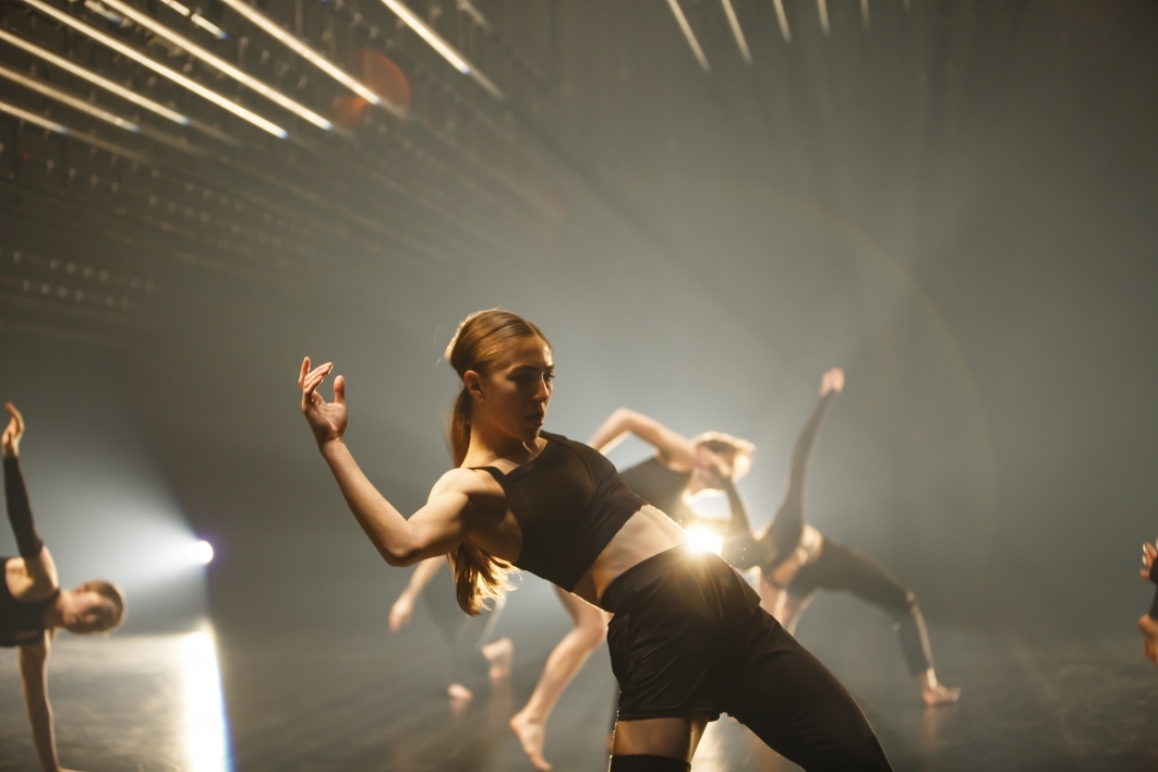 Dancer mid-performance wearing all black with other dancers and lights in the background.