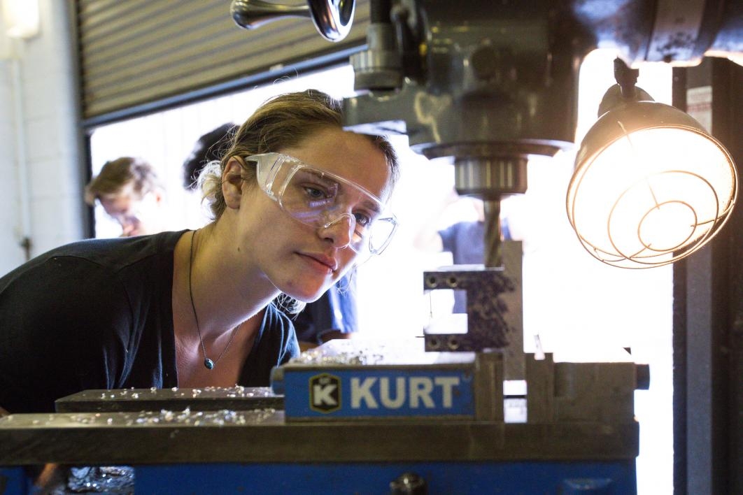 A woman watches the milling of a piece of metal on a drill machine