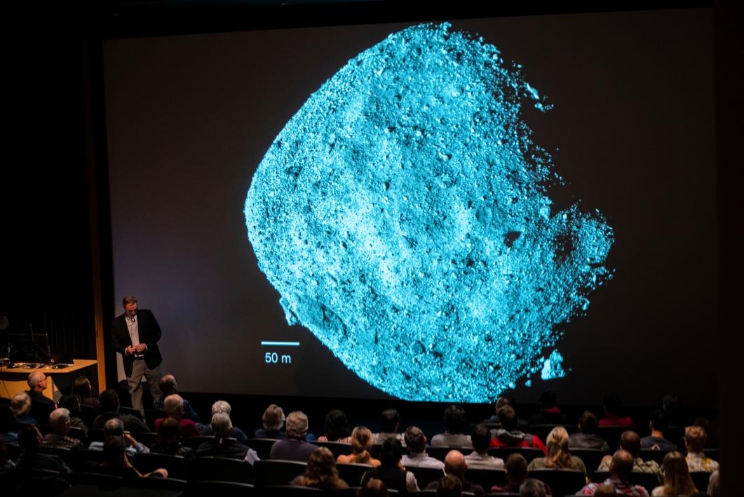 A screen shows the asteroid Bennu to an audience