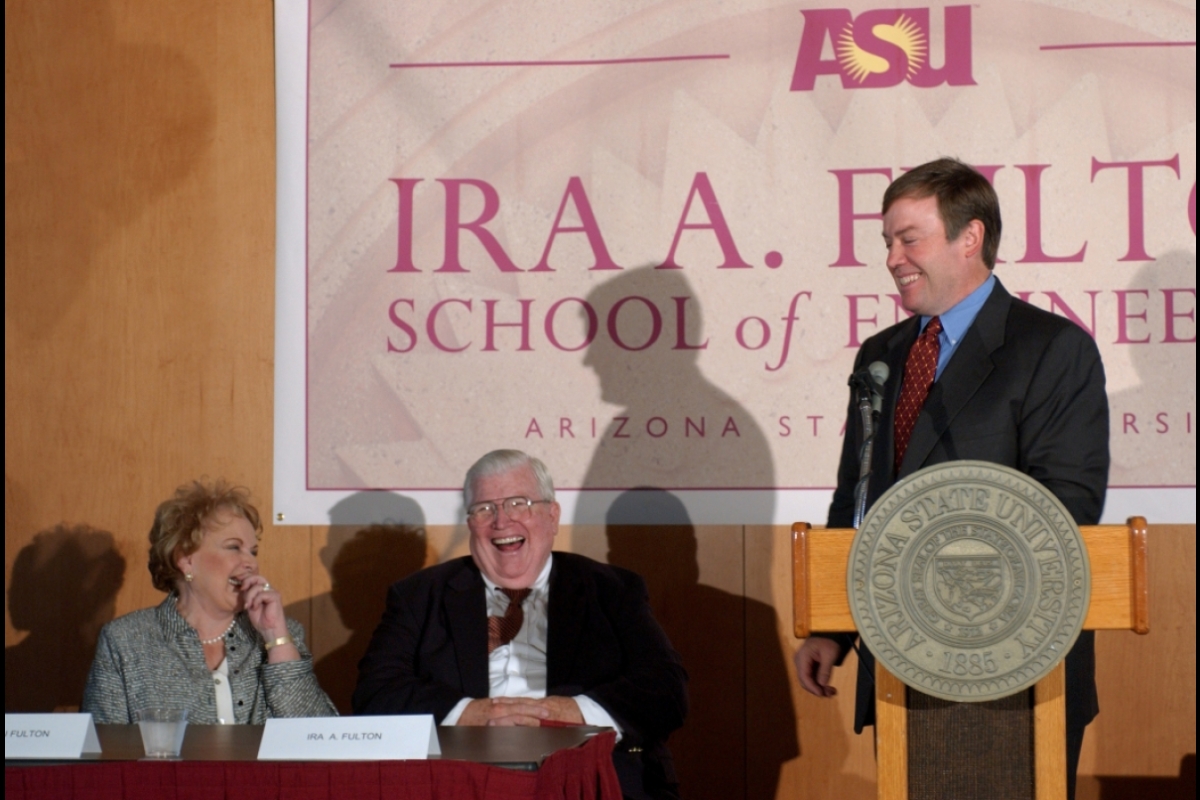 Man speaking behind a lectern with the Arizona State University seal on it as a seated man and woman listen and laugh.