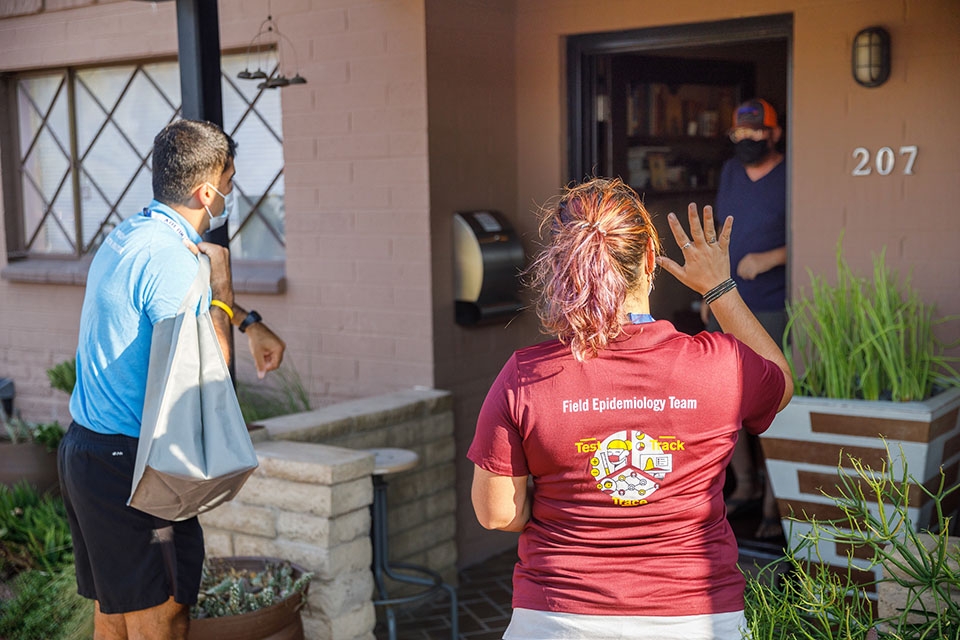 Two volunteers, in blue and maroon shirts, greet a man opening the door to his home.
