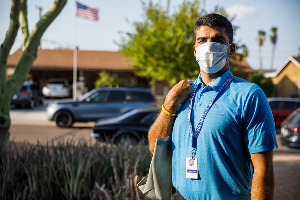 A man wearing a protective mask, a blue shirt and a Maricopa County Department of Public Health volunteer lanyard stands in an Arizona neighborhood.
