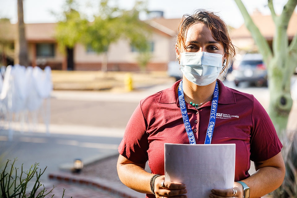 A woman wearing a protective mask, a maroon shirt and a blue lanyard stands in an Arizona neighborhood.