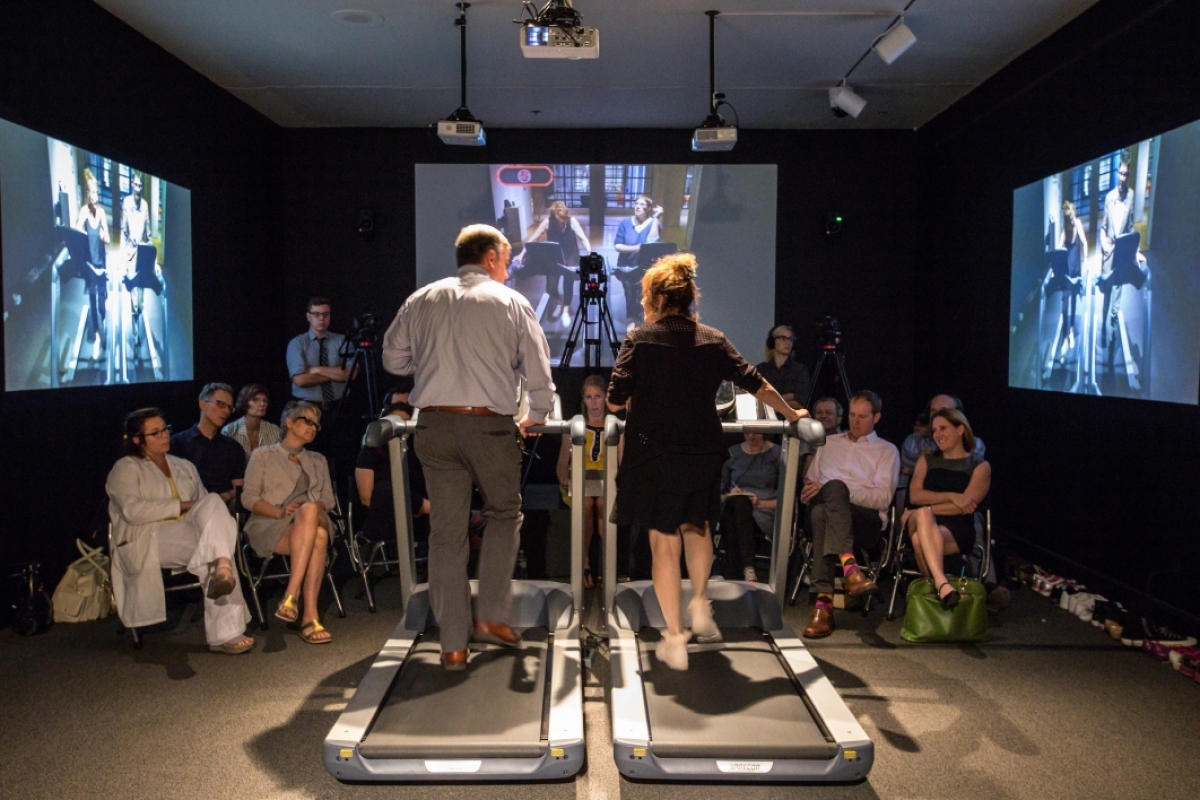 Mand and woman seen from behind, each standing on a treadmill while speaking, as a crowd of seated onlookers watch.