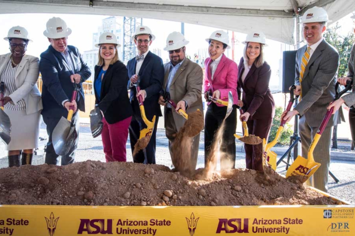 A line of people in suits and hard hats turn ceremonial dirt with shovels at a groundbreaking