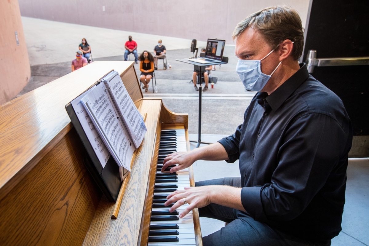 An opera teacher plays an upright piano as his students practice in a loading dock