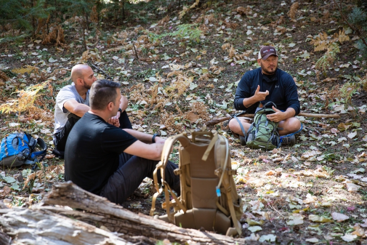 A few men seated on a leaf-covered ground area outdoors, talking.
