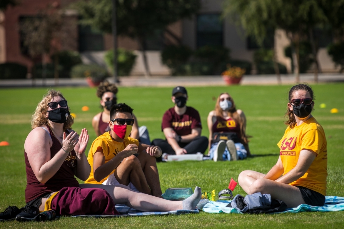 Small groups of students sit spaced out on a field to watch the ASU football game on a giant TV