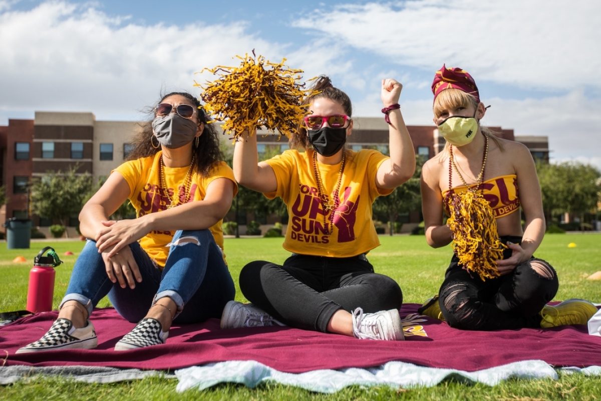 Three students in ASU gear cheer at a game watching party outside at the West campus