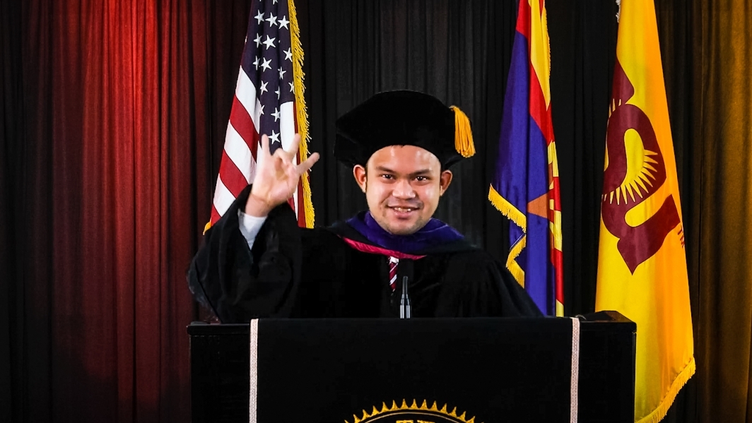 A graduate speaks at a lectern in front of flags