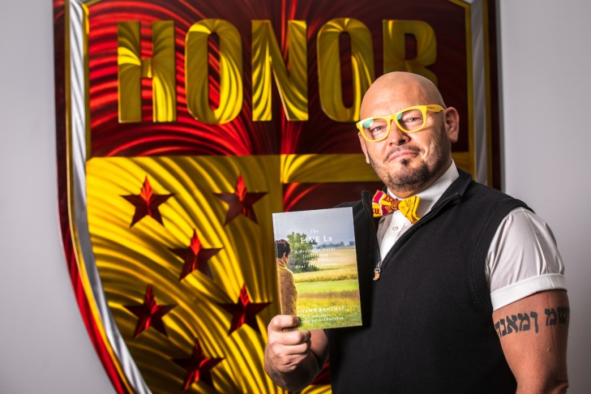 Man wearing glasses and a bow tie holds a book in front of a large shield that says "honor."