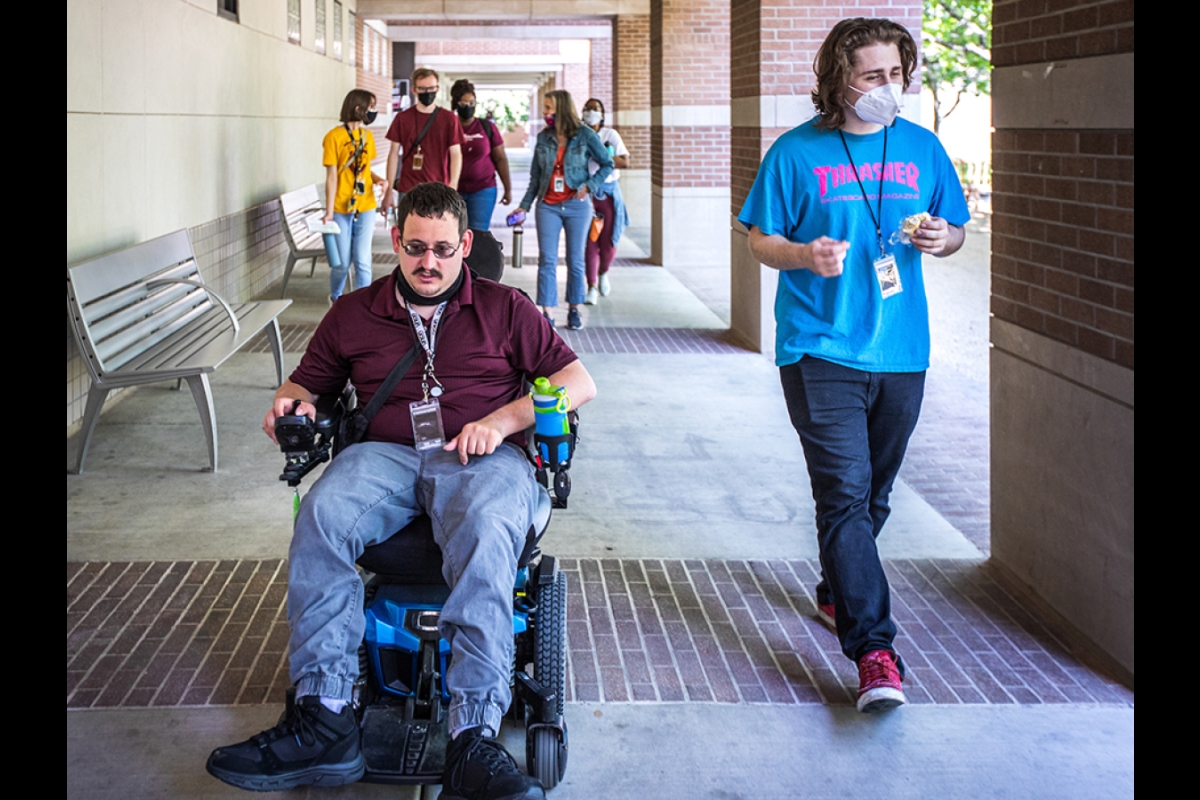man in wheelchair and man walking leading a group behind them down a campus corridor