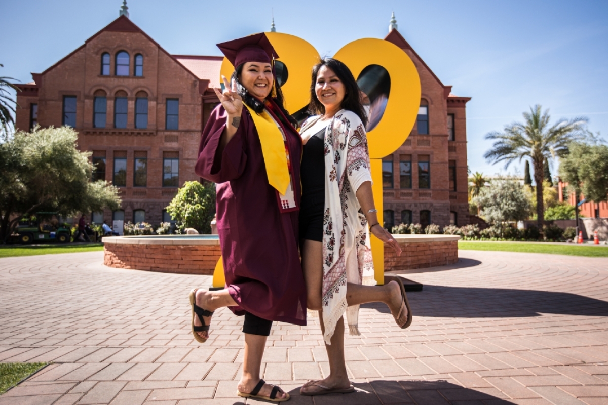 A woman in a graduation cap and gown poses with her sister in front of a giant 2021 sign