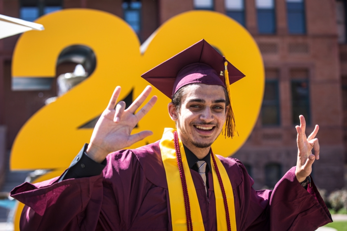 A man in a graduation cap and gown flashes the pitchfork gesture and smiles for a photo in front of a giant 2021 sign