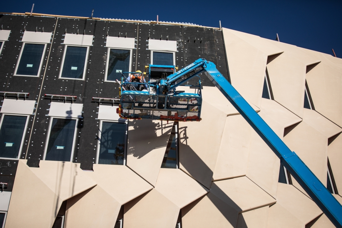 Two men in a lift inspect concrete panels being installed on the side of a building