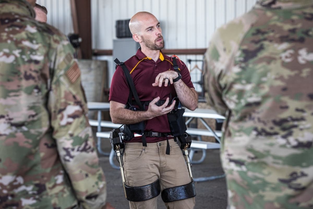 A man not in uniform demonstrates an exoskeleton to a group in military camo