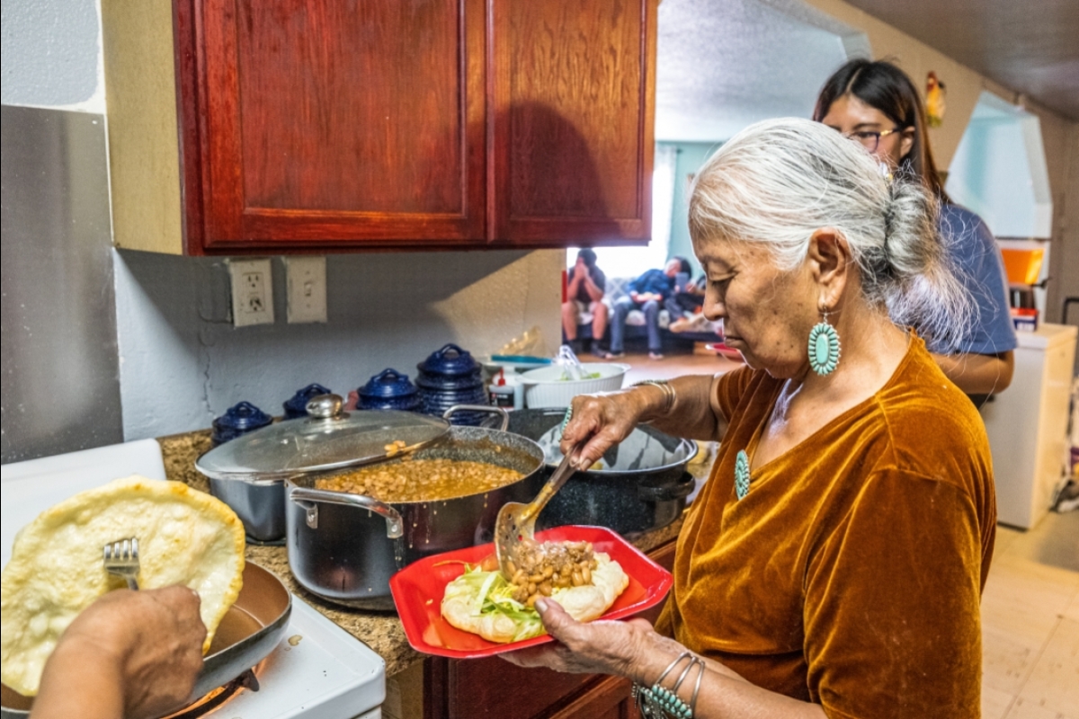 An older Navajo woman scoops beans onto a piece of frybread as she prepares a Navajo taco in a kitchen